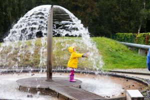 Kind in Regenmantel steht unter dem Wasserstrahl eines Brunnens