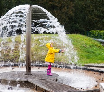 Kind in Regenmantel steht unter dem Wasserstrahl eines Brunnens
