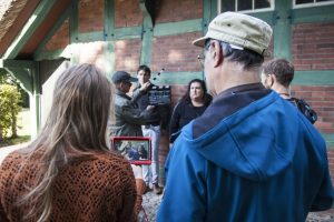 Regisseur:innen schauen auf Bildschirm, gleichzeitig hält Person Filmklappe vor die Szene