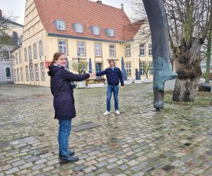zwei Personen reichen sich die Hand und stehen vor Haus auf Kopfsteinpflaster