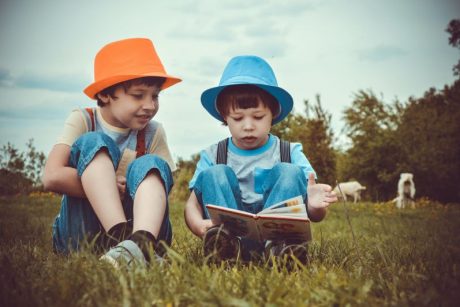 Zwei Kinder mit Hüten sitzen draußen im Gras und lesen ein Buch