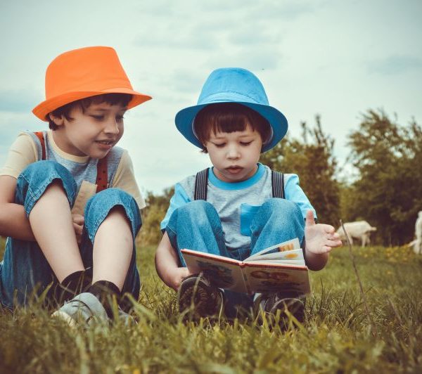 Zwei Kinder mit Hüten sitzen draußen im Gras und lesen ein Buch