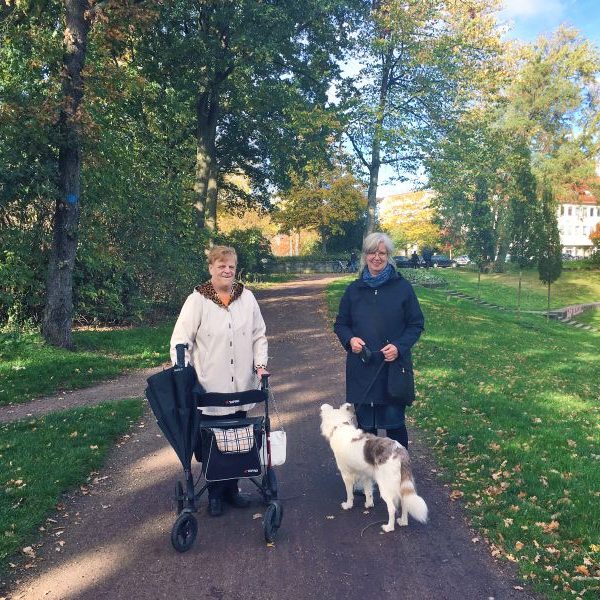 Zwei Frauen gehen spazieren. Eine mit Rollator, die andere mit einem Hund.
