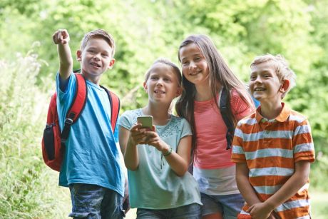 Vier lachende Kinder stehen im Freien auf einem Wanderweg, zwei mit Rucksäcken. Eines hält ein Smartphone, ein anderes zeigt nach vorn. Im Hintergrund ist grüne Natur zu sehen.