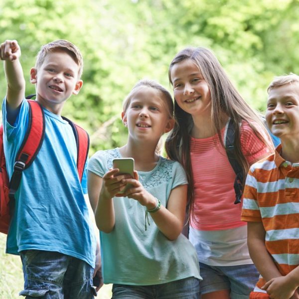 Vier lachende Kinder stehen im Freien auf einem Wanderweg, zwei mit Rucksäcken. Eines hält ein Smartphone, ein anderes zeigt nach vorn. Im Hintergrund ist grüne Natur zu sehen.
