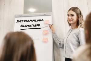 Eine junge Frau steht lächelnd neben einem Flipchart und zeigt mit einem Stift auf mehrere Haftnotizen. Auf dem Flipchart steht in großer Schrift „Ideensammlung Klimatreff“. Im Vordergrund sind verschwommen die Köpfe von Zuhörer:innen zu sehen. Die Szene findet in einem modernen, hellen Raum statt, vermutlich während eines Workshops oder Meetings zum Thema Klima. Eine junge Frau steht lächelnd neben einem Flipchart und zeigt mit einem Stift auf mehrere Haftnotizen. Auf dem Flipchart steht in großer Schrift „Ideensammlung Klimatreff“. Im Vordergrund sind verschwommen die Köpfe von Zuhörer:innen zu sehen. Die Szene findet in einem modernen, hellen Raum statt, vermutlich während eines Workshops oder Meetings zum Thema Klima. 
